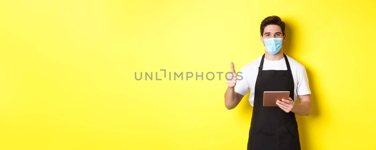 Concept of covid-19, small business and pandemic. Friendly waiter in medical mask and black apron showing thumb up, taking orders with digital tablet, yellow background by Benzoix
