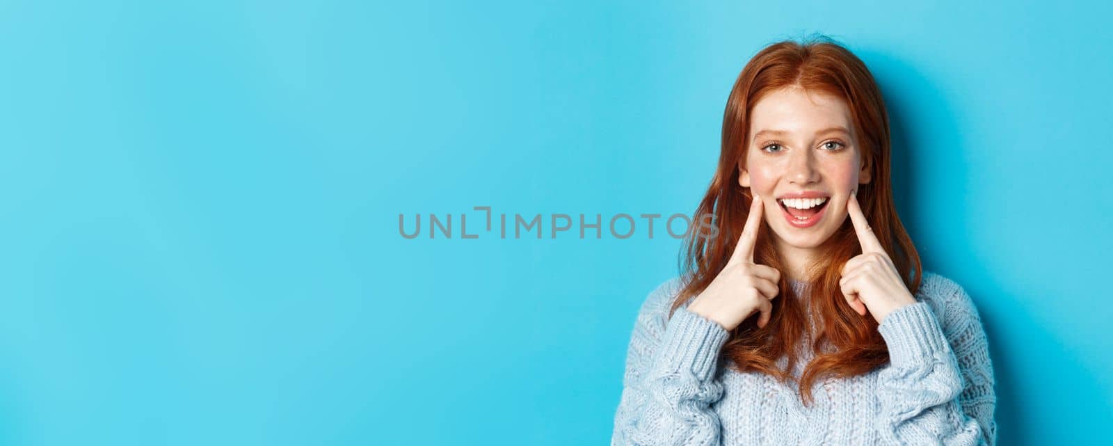 Close-up of cheerful teenage girl with red hair and freckles, poking cheeks, showing dimples and smiling with white teeth, standing over blue background by Benzoix