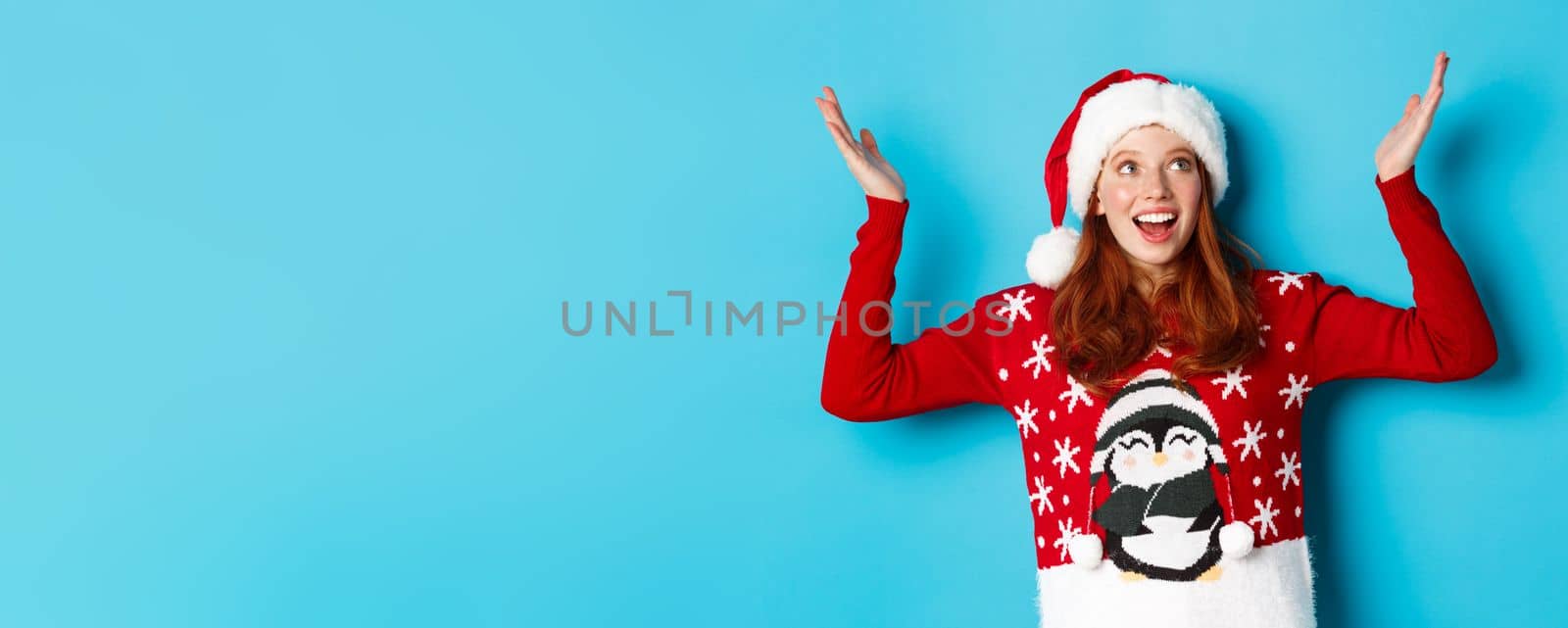 Happy holidays and Christmas concept. Excited redhead girl rejoicing of something falling on her, raising hands up delighted and smiling, wearing Santa hat with xmas sweater, blue background.