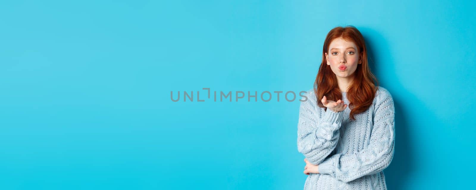 Lovely teen girl in sweater blowing air kiss, pucker lips and staring at camera, standing against blue background by Benzoix