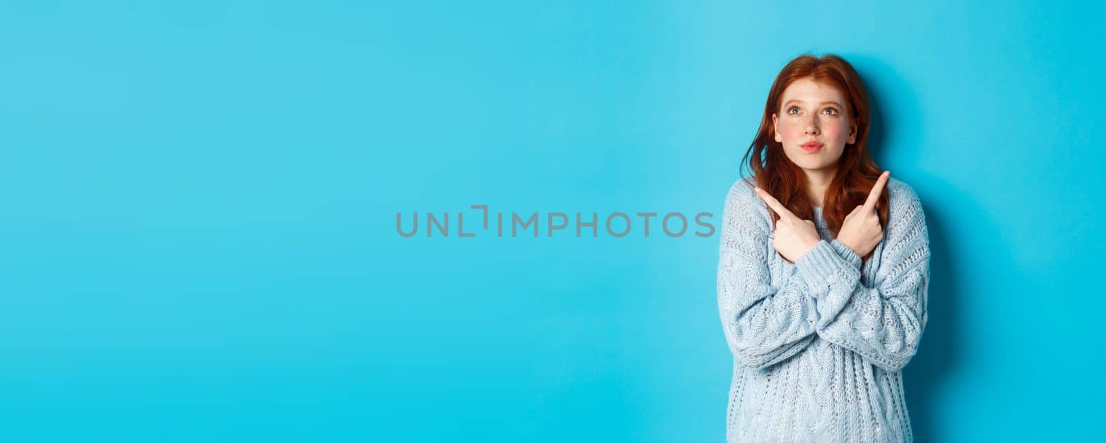 Excited redhead girl pointing fingers sideways, showing two choices and looking tempted up, standing against blue background.