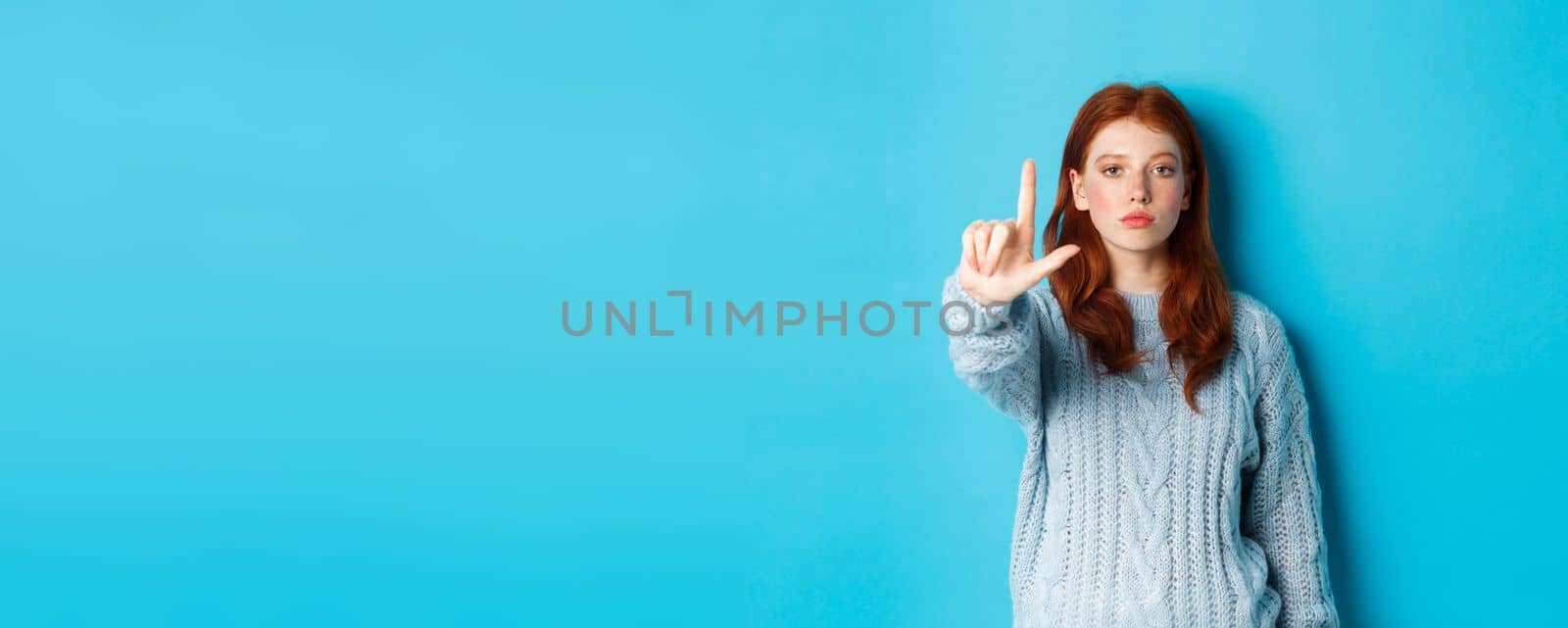 Serious redhead girl in sweater showing taboo gesture, extending one finger, shaking forefinger to disapprove, disagree and forbid something, blue background by Benzoix