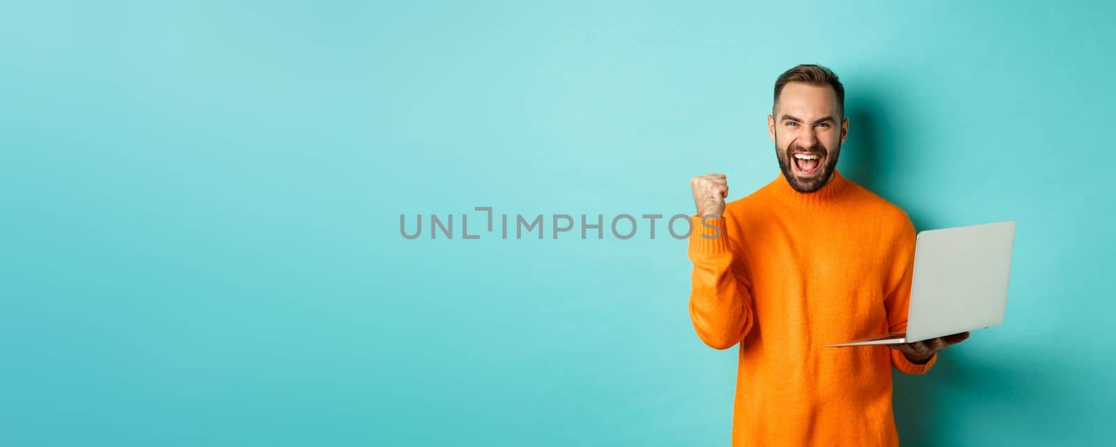 Freelance and technology concept. Lucky man winner celebrating, winning online, showing fist pump and holding laptop, standing over light blue background.
