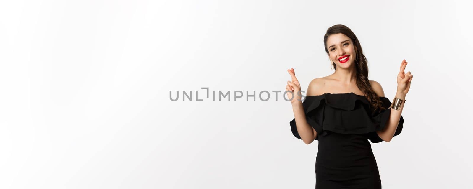 Fashion and beauty. Stylish glamour woman in black dress, red lips, looking optimistic and smiling while cross fingers, making wish, standing over white background by Benzoix