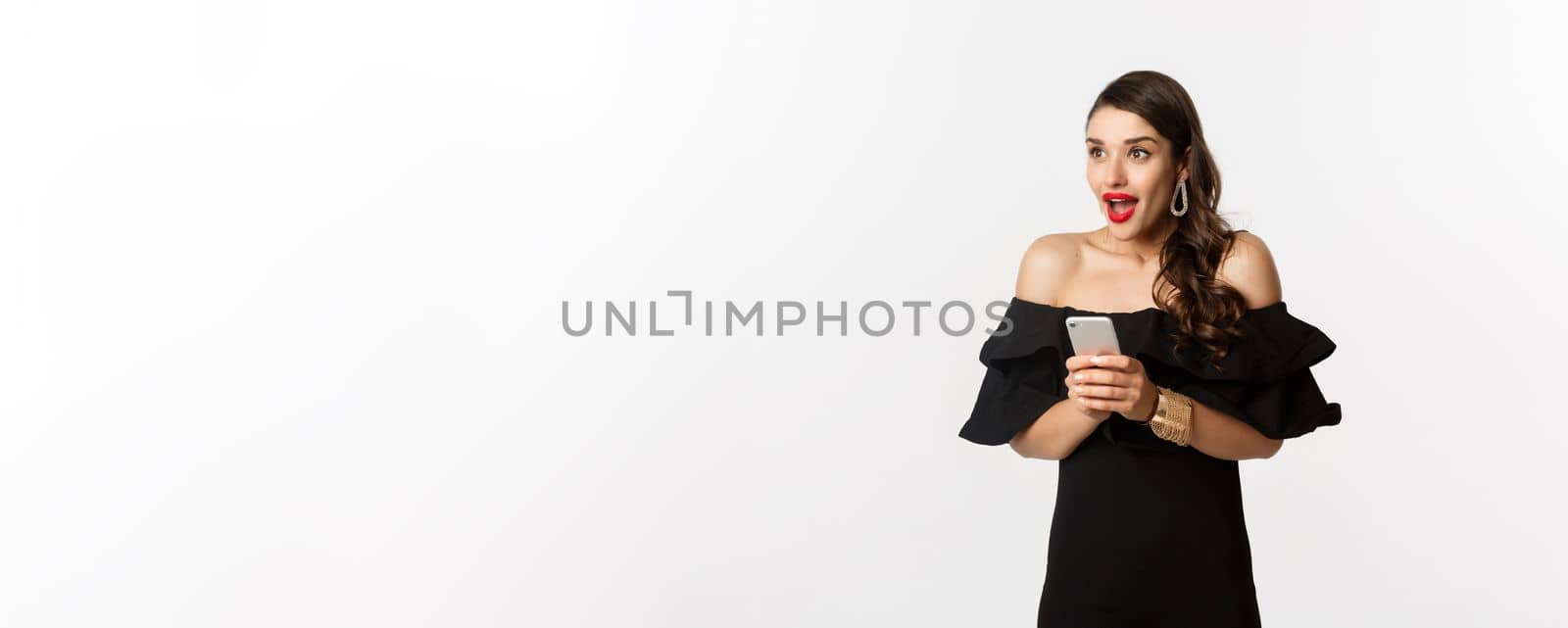 Image of excited woman in black glamour dress, red lipstick, using mobile phone and looking left amazed, standing over white background.