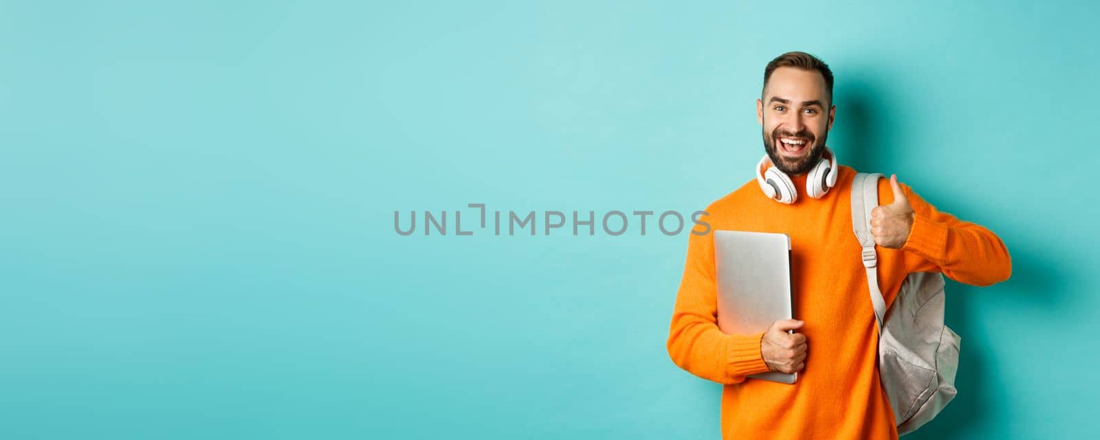 Happy man with backpack and headphones, holding laptop and smiling, showing thumb-up in approval, standing over turquoise background by Benzoix