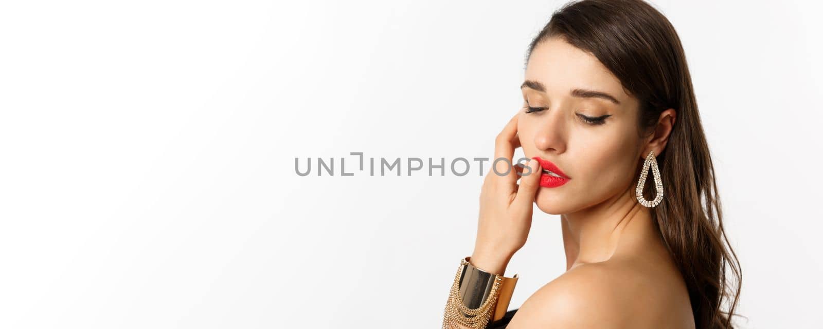 Fashion and beauty concept. Tender and feminine brunette woman touching red lips and looking down sensually, standing over white background.