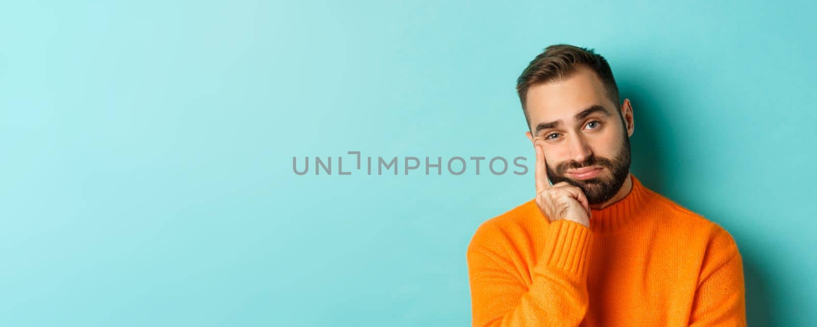 Close-up of unamused sad man looking at camera, pouting and leaning face on hand, standing over light blue background by Benzoix