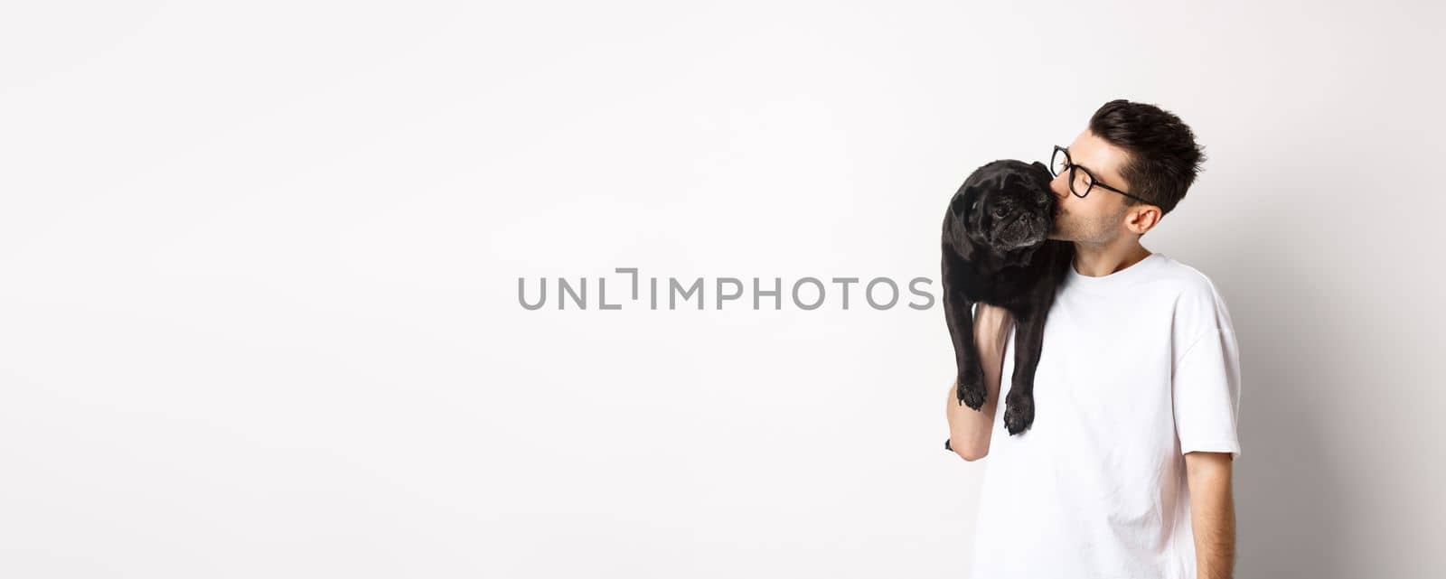 Image of handsome young man kissing his cute black pug, holding dog on shoulder, standing over white background.