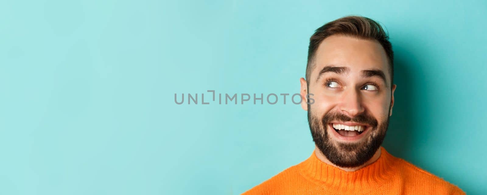 Headshot of handsome caucasian man looking at upper left corner logo and smiling pleased, checking out promo offer, standing over turquoise background by Benzoix