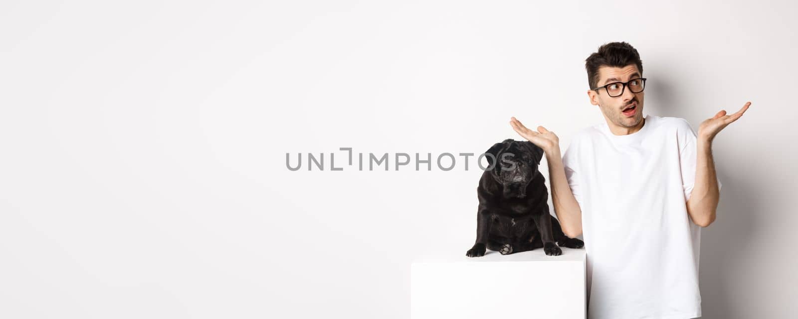 Portrait of confused hipster guy dog owner shrugging, standing near cute black pug pet, white background.