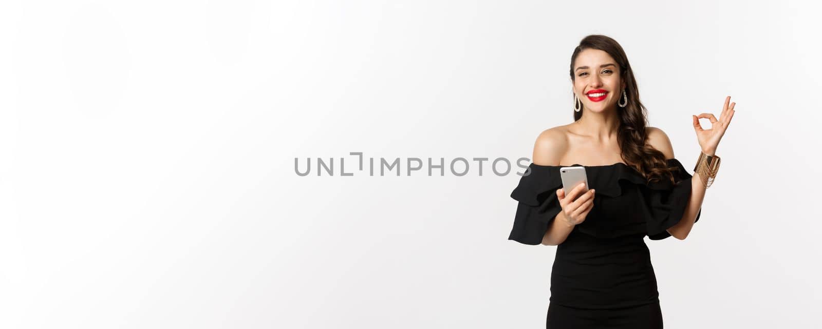 Online shopping concept. Attractive woman in trendy black dress, makeup, showing okay sign in approval and using mobile phone app, white background by Benzoix