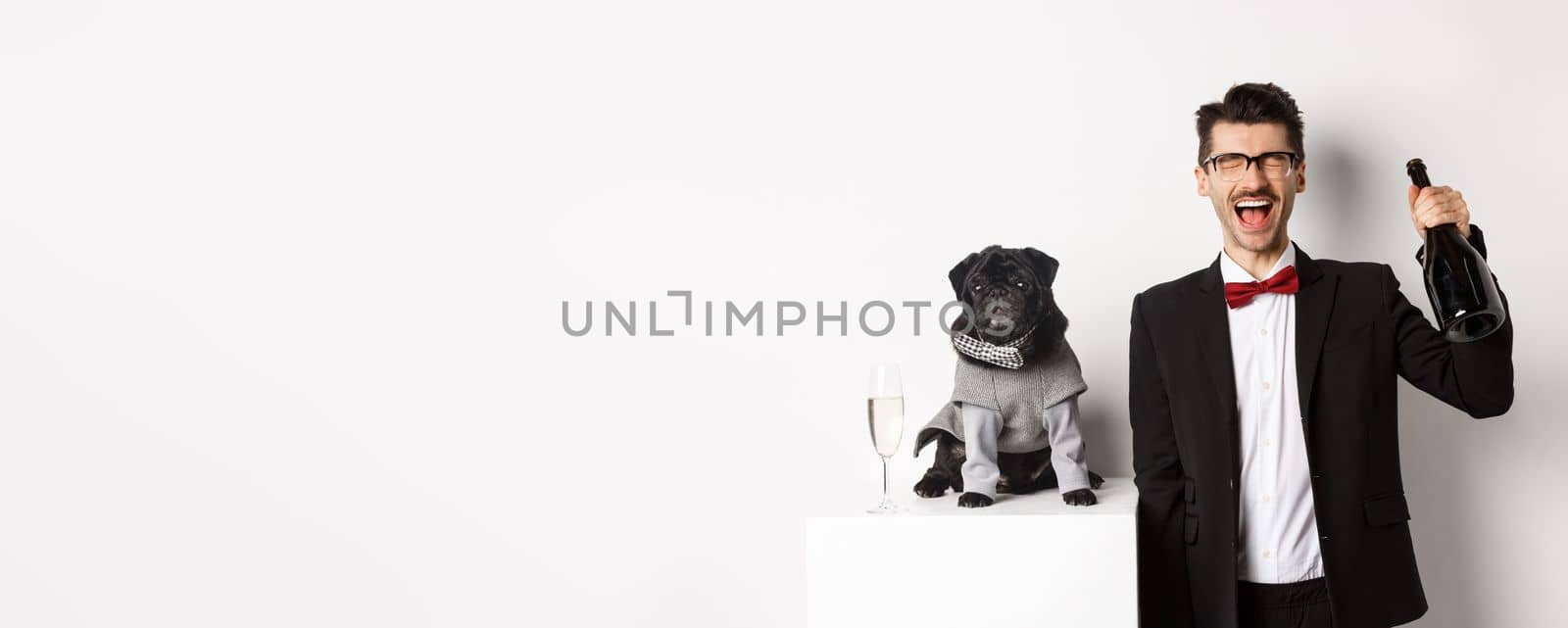 Pets, winter holidays and New Year concept. Happy man celebrating Christmas party pet, standing with cute dog in costume, drinking champagne and rejoicing, white background.