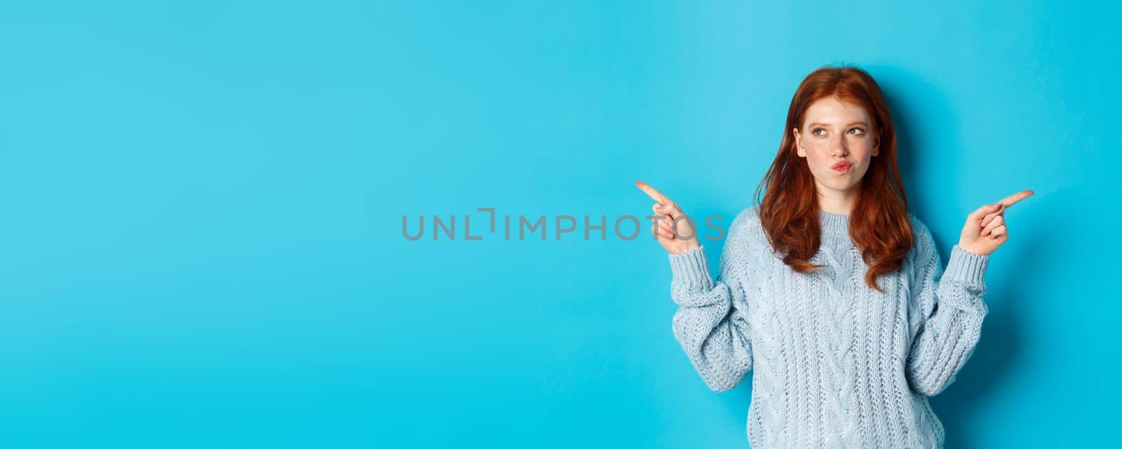 Winter holidays and people concept. Thoughtful redhead girl making decision, pointing fingers sideways, choosing between two ways, standing over blue background.