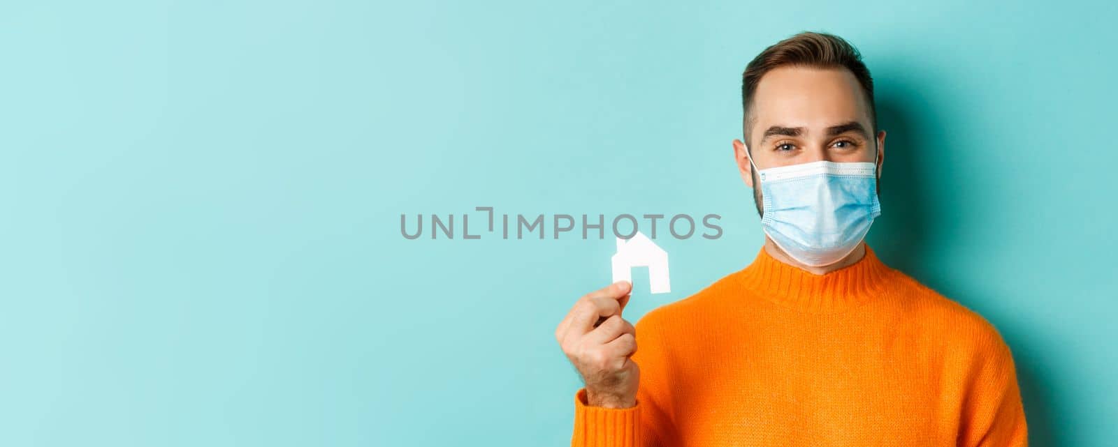 Real estate and coronavirus pandemic concept. Close-up of adult man in medical mask holding small paper house maket and smiling, searching for apartment, light blue background.