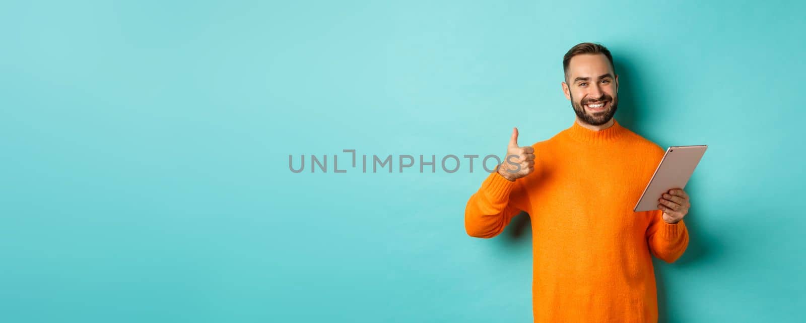 Satisfied adult man smiling, using digital tablet and showing thumb-up, approve and agree, standing against turquoise background by Benzoix