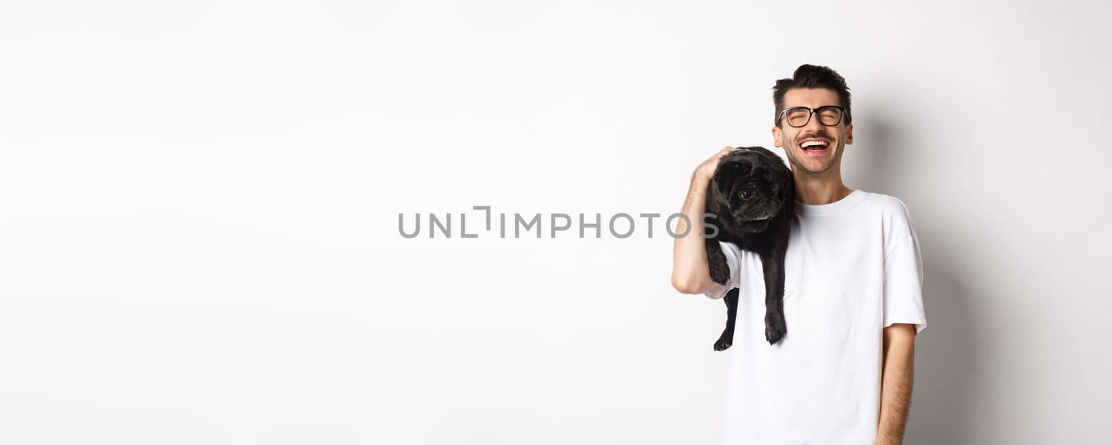 Happy young man laughing carefree, holding cute black dog, pug breed, on shoulder and having fun, standing over white background by Benzoix