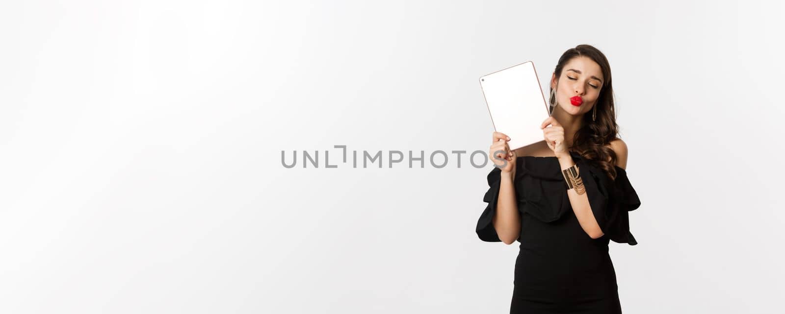Fashion and shopping concept. Beautiful woman with red lips, wearing black dress, showing digital tablet and making kissing face, white background.