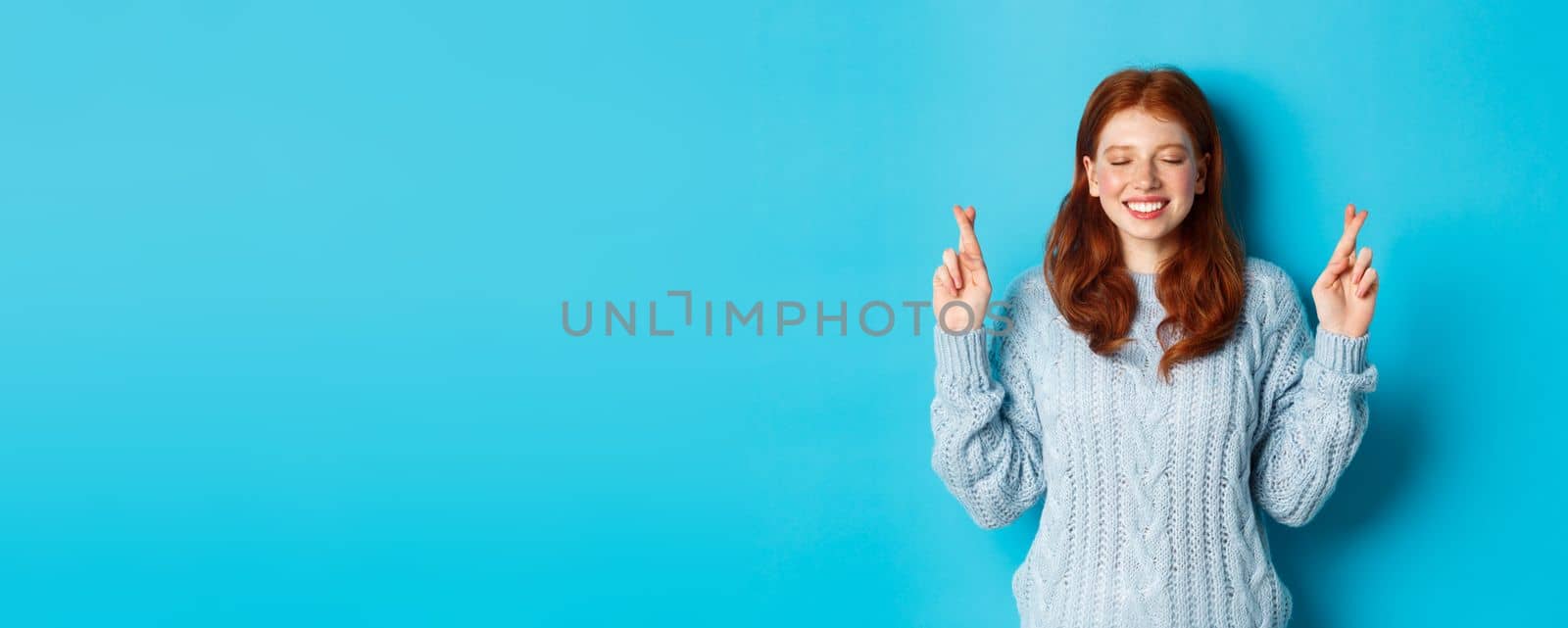 Hopeful redhead girl making a wish, cross fingers for good luck, smiling and anticipating good news or positive result, standing against blue background.