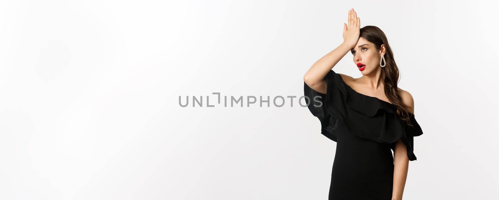 Fashion and beauty. Annoyed stylish woman in black dress, slap forehead, making facepalm and roll eyes tired, standing over white background.
