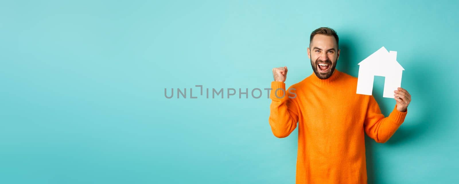 Real estate concept. Excited man saying yes, showing paper house maket and looking satisfied, standing in orange sweater over light blue background.