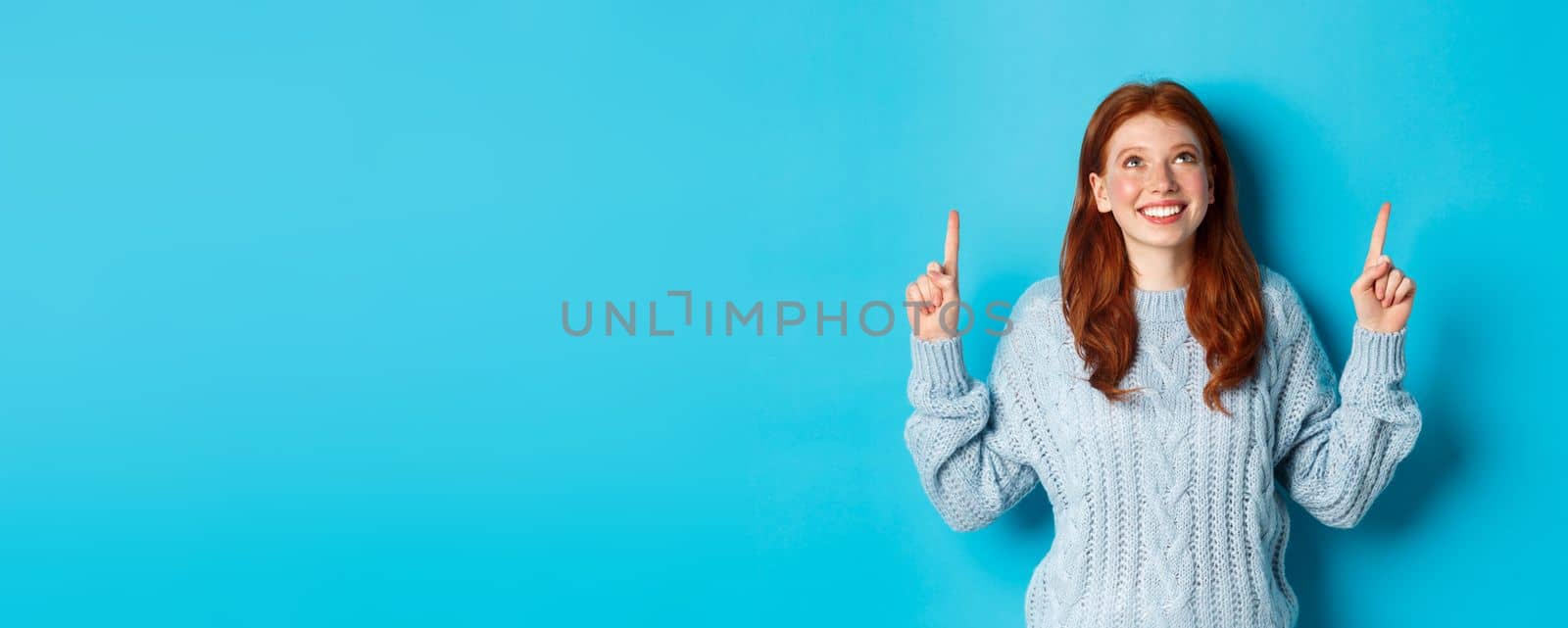Winter holidays and people concept. Cute teenage redhead girl pointing fingers up, looking at top promo and smiling amused, standing over blue background.