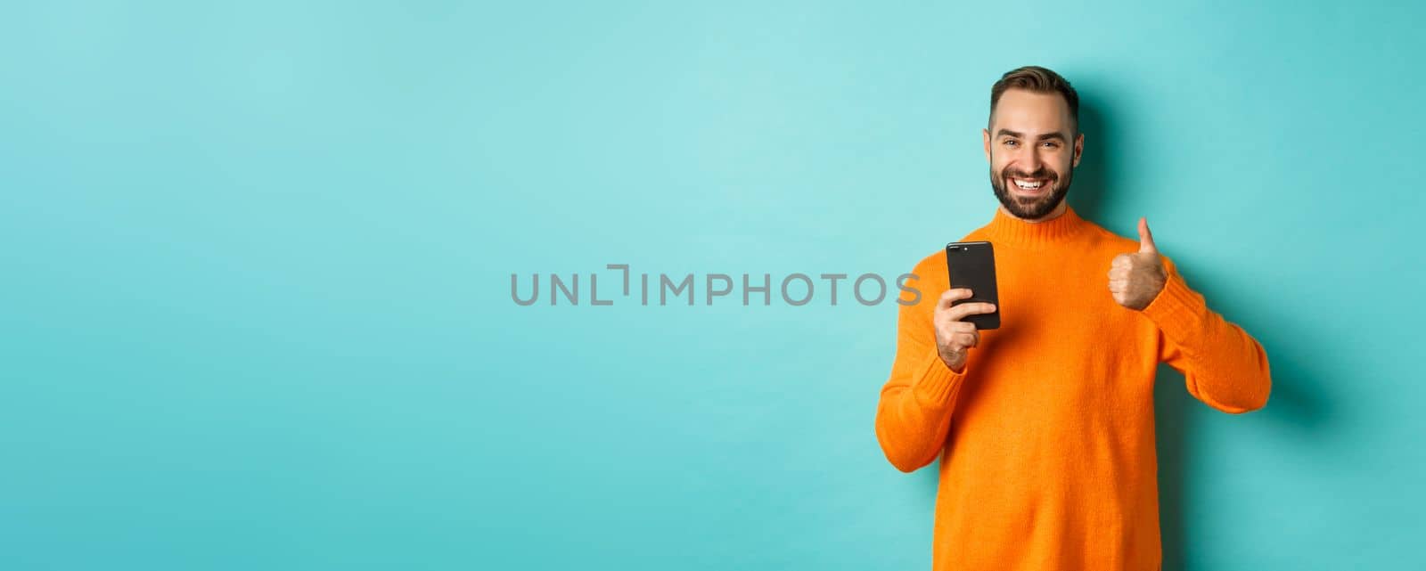 Photo of satisfied young man in orange sweater, showing thumb-up after reading on mobile phone, standing pleased against turquoise background.