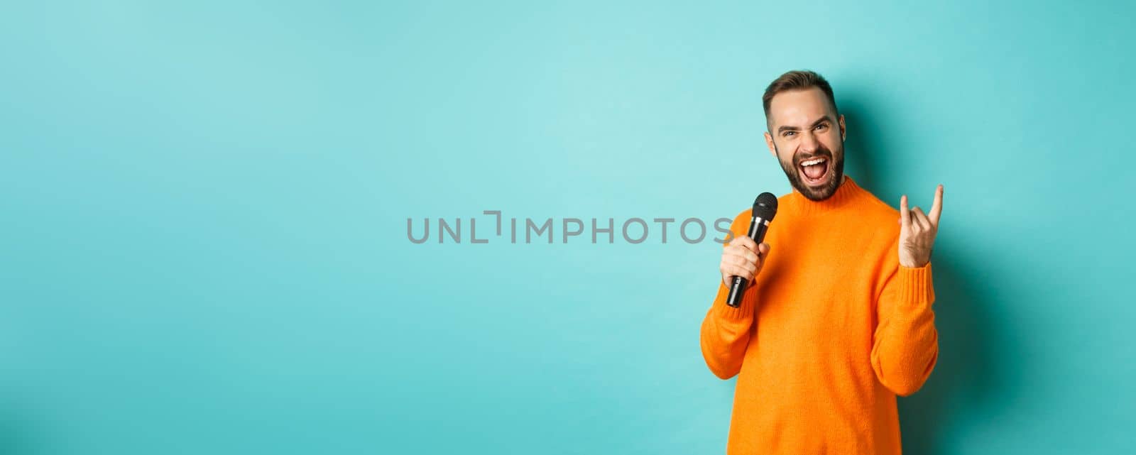 Joyful handsome man singing song in microphone, showing rock-n-roll gesture excited, standing over turquoise background.