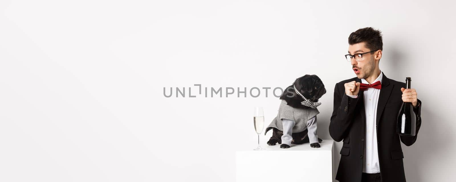 Pets, winter holidays and New Year concept. Happy young man celebrating Christmas with cute black dog wearing party costume, puppy looking at owner, white background.