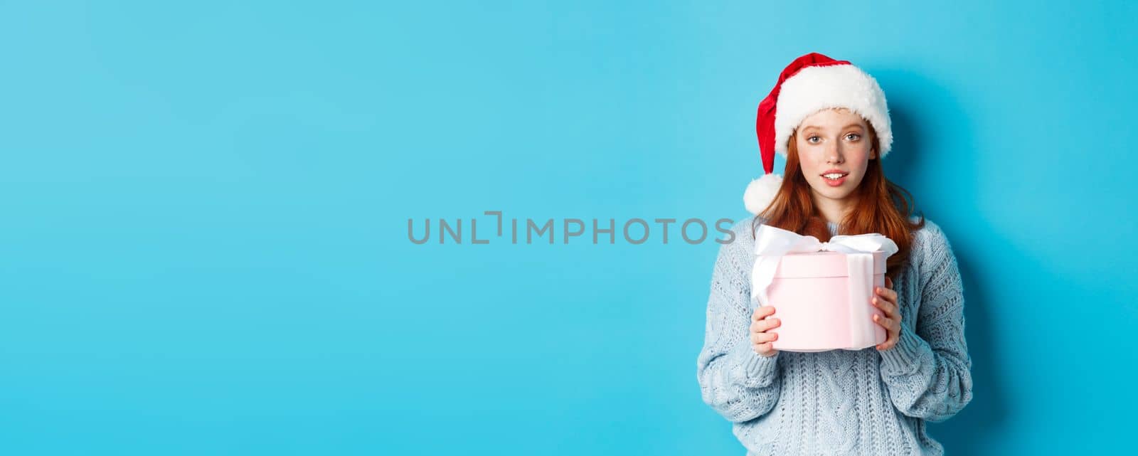 Winter holidays and Christmas Eve concept. Cute redhead girl wearing Santa hat, holding New Year gift and looking at camera, standing against blue background by Benzoix