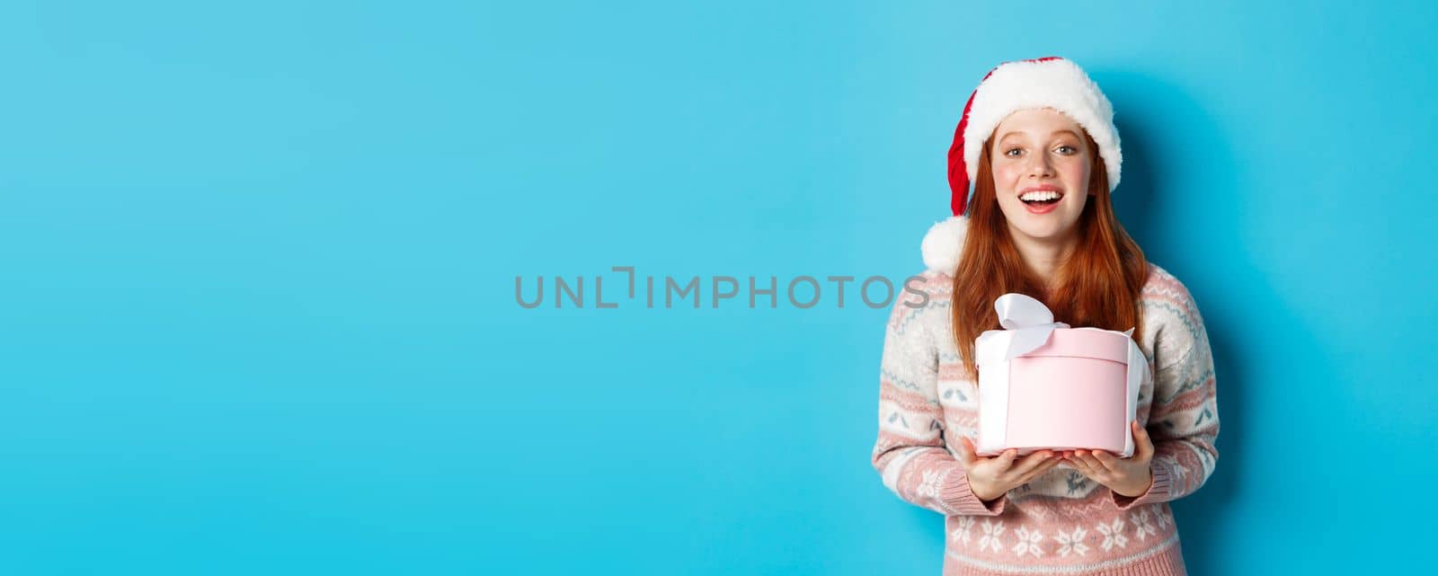 Winter and celebration concept. Happy redhead girl receiving christmas gift and thanking, hugging box with presents and smiling, standing in santa hat over blue background by Benzoix