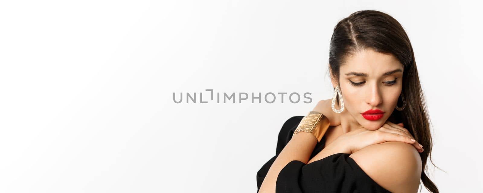 Fashion and beauty concept. Close-up of sensual woman in elegant earrings and black dress, wearing makeup with red lips, looking down tenderly, standing over white background.