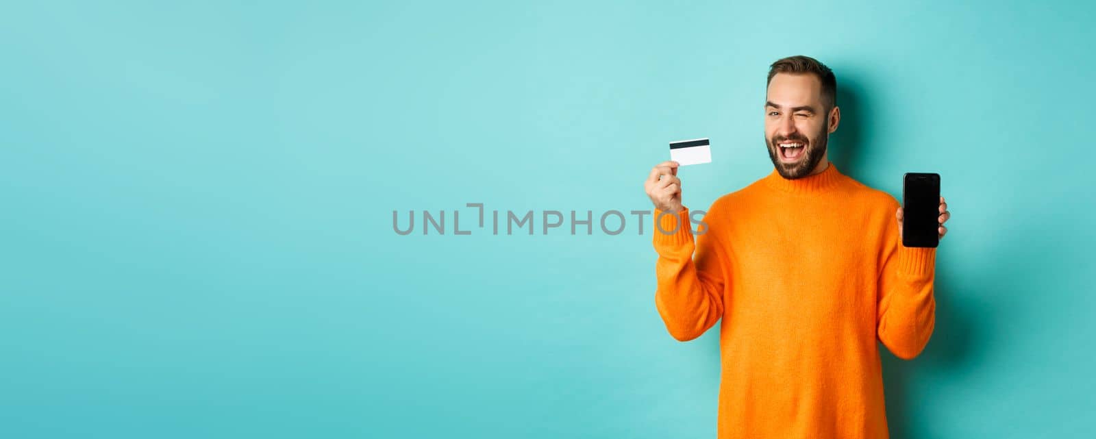 Online shopping. Handsome caucasian man showing credit card and smartphone, winking at camera, recommending something, standing over light blue background.