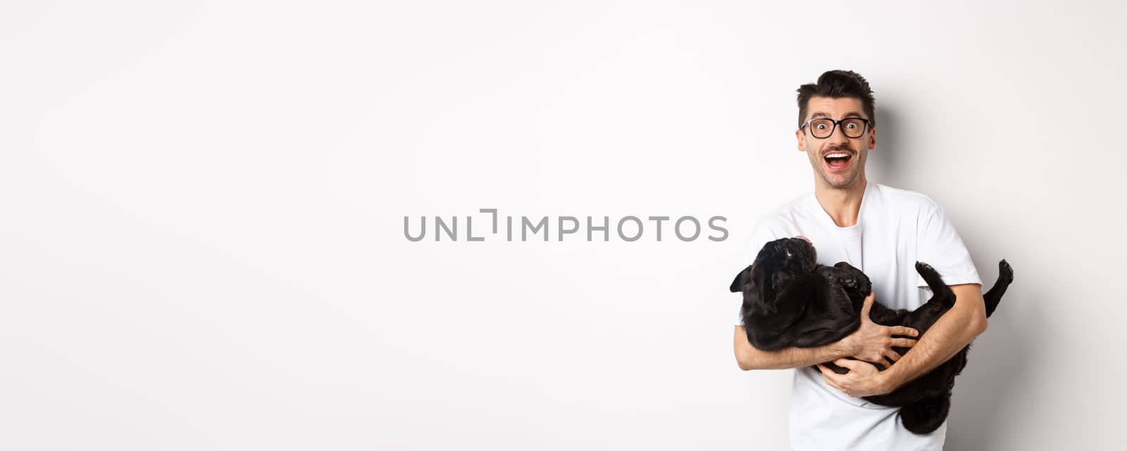 Handsome young man playing with cute black puppy. Dog owner petting a pug, standing over white background.