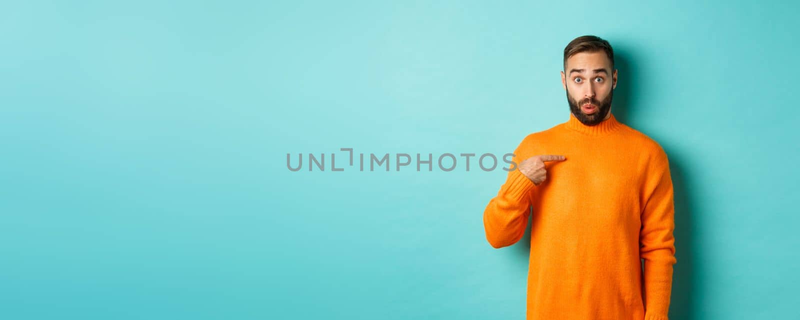 Man pointing at himself with surprise face, being chosen, standing confused against light blue background.