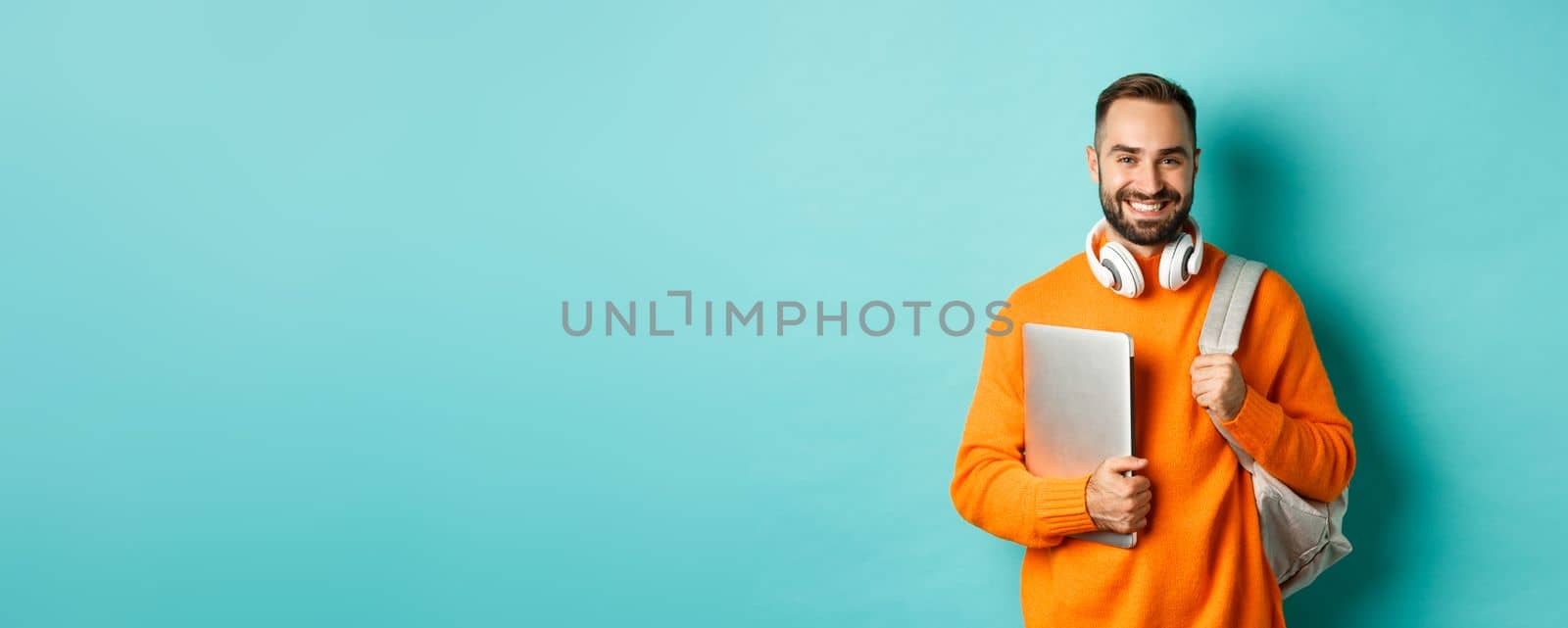 Happy man with backpack and headphones, holding laptop and smiling, going to work, standing over turquoise background.