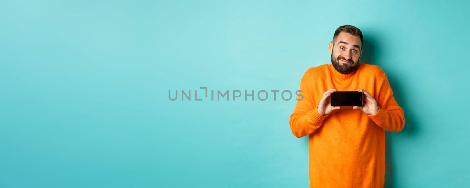 Clueless guy shrugging and showing mobile screen, indecisive emotion, standing in orange sweater over turquoise background.