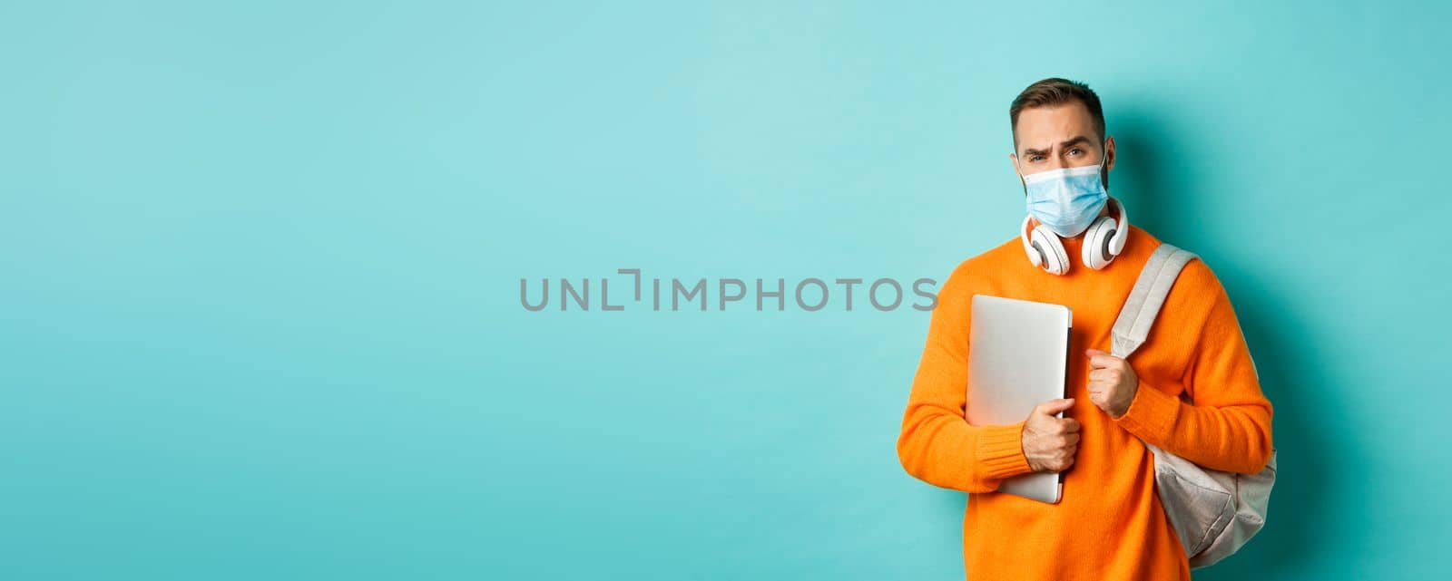 Handsome caucasian man with headphones and backpack, holding laptop and wearing medical mask, looking doubtful and skeptical, standing over light blue background.