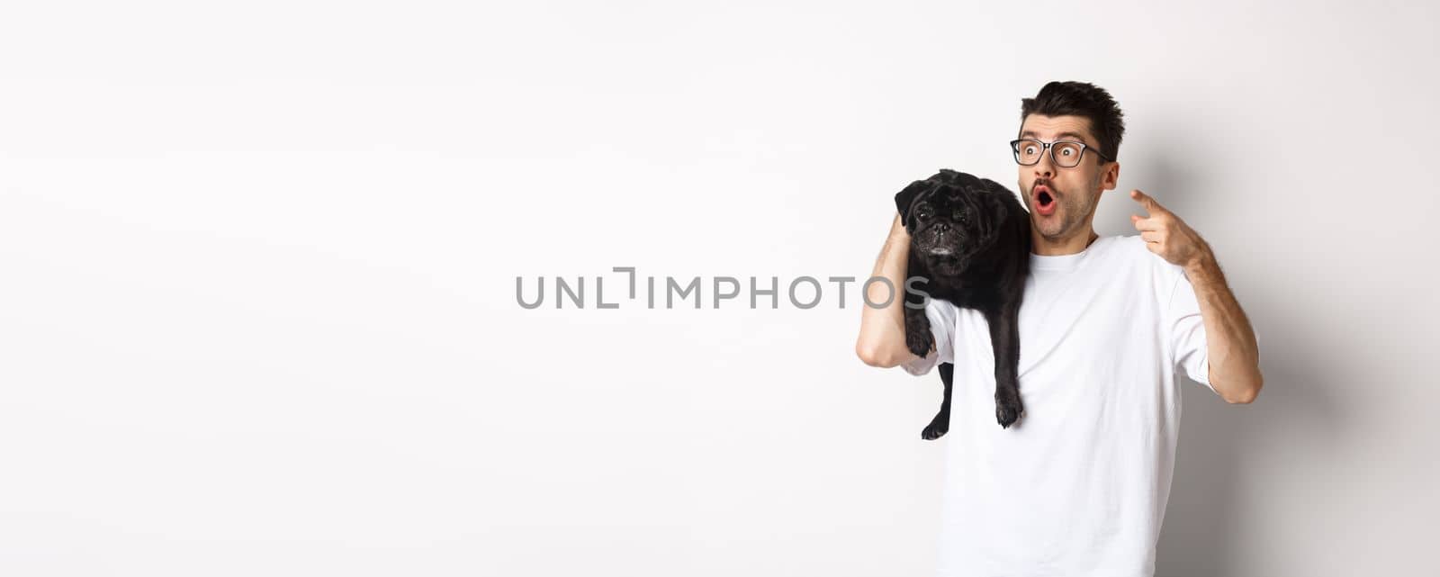 Amazed young man holding cute black dog on shoulder, pointing finger left at promo offer, staring impressed and speechless, standing over white background.