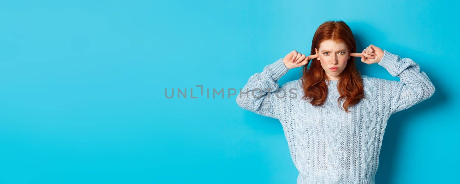 Teenage redhead girl unwilling to listen, shut ears and frowning angry, staring at camera offended, sulking against blue background.