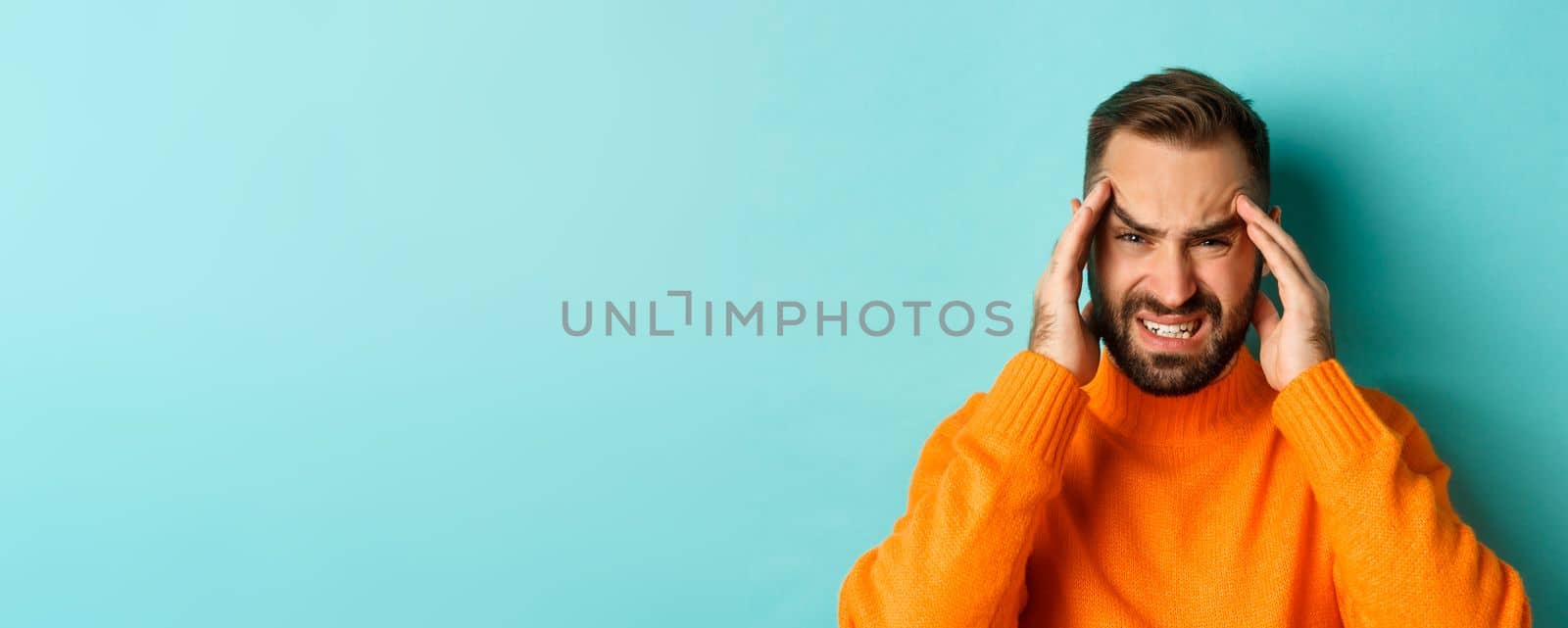 Close-up of troubled man having headache, grimacing and touching head, suffering migraine, standing over light blue background.