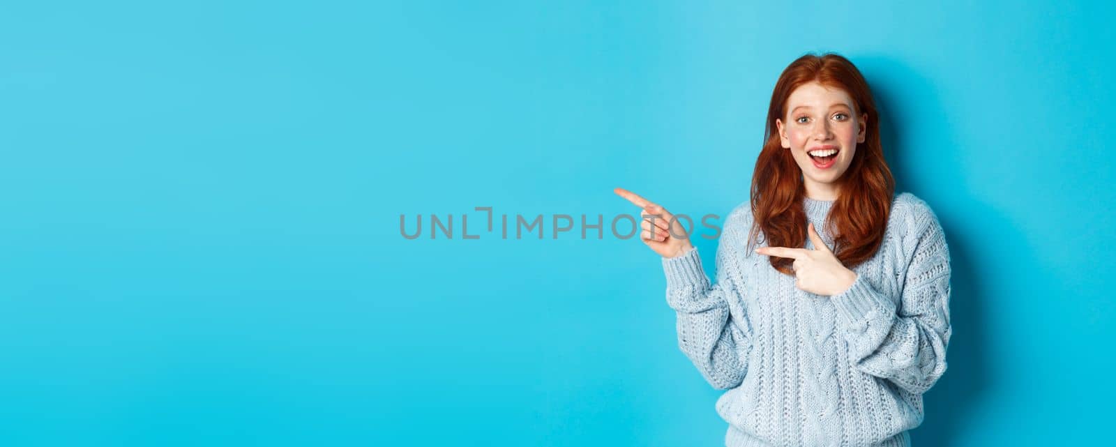 Amazed teenage girl with red hair and freckles, pointing fingers left at logo and smiling, showing advertisement, standing over blue background.