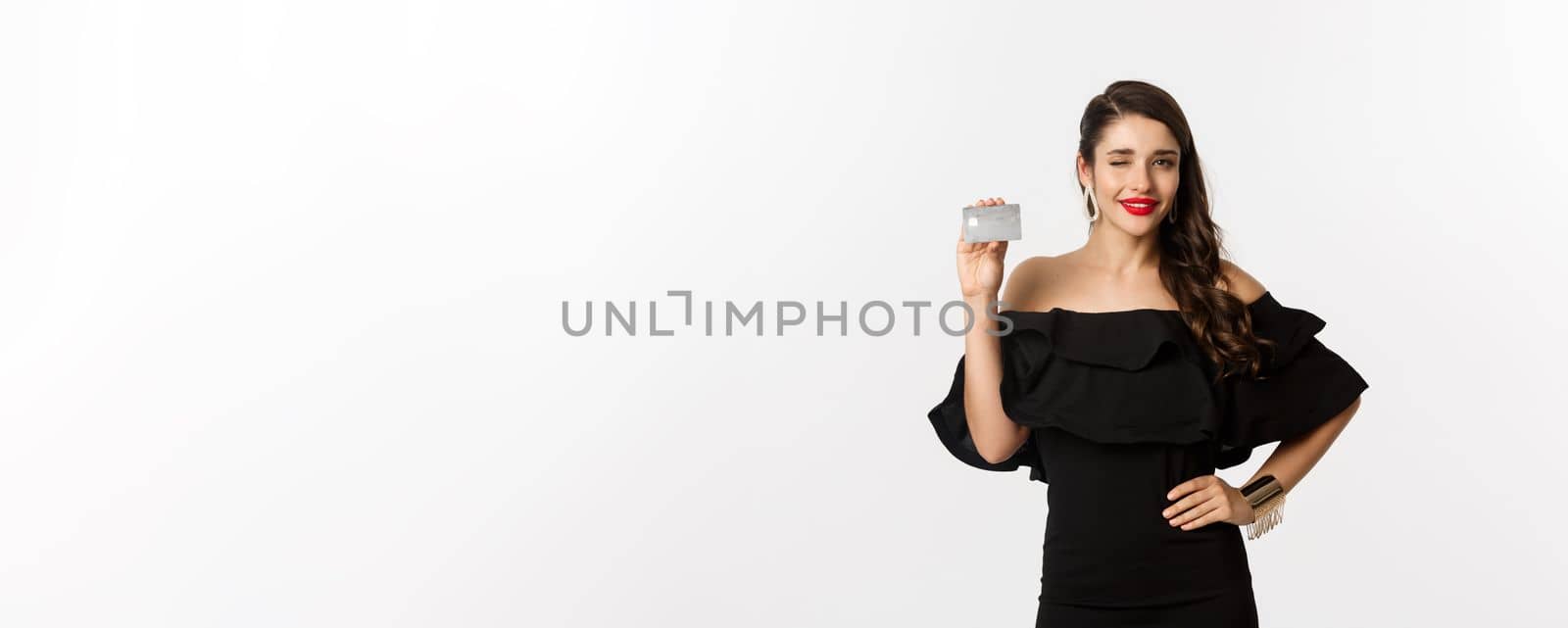 Beauty and shopping concept. Fashionable woman in black dress, wearing makeup, winking at camera and showing credit card, standing over white background.