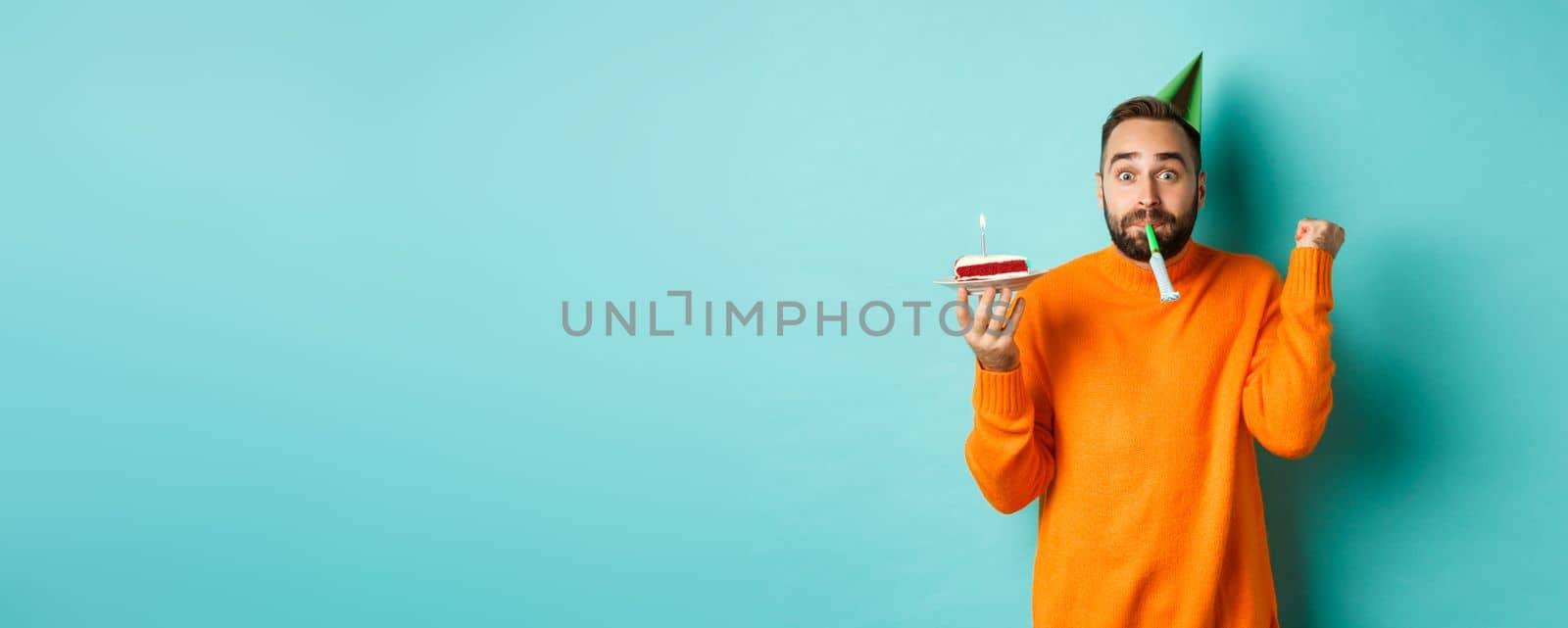 Happy birthday guy celebrating, wearing party hat, blowing wistle and holding bday cake and doing fist pump excited, standing over turquoise background.