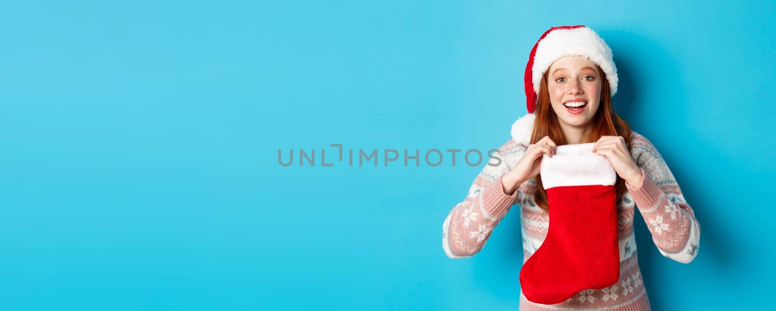 Beautiful redhead girl in santa hat open Christmas stocking and looking surprised, receiving xmas gift, standing over blue background.