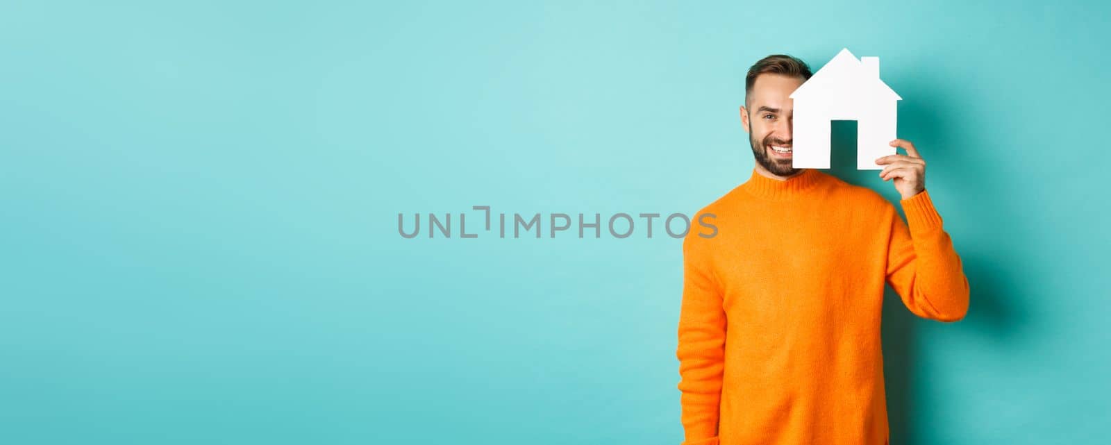 Real estate concept. Happy young man searching for home rent, holding house paper maket and smiling, standing over blue background.