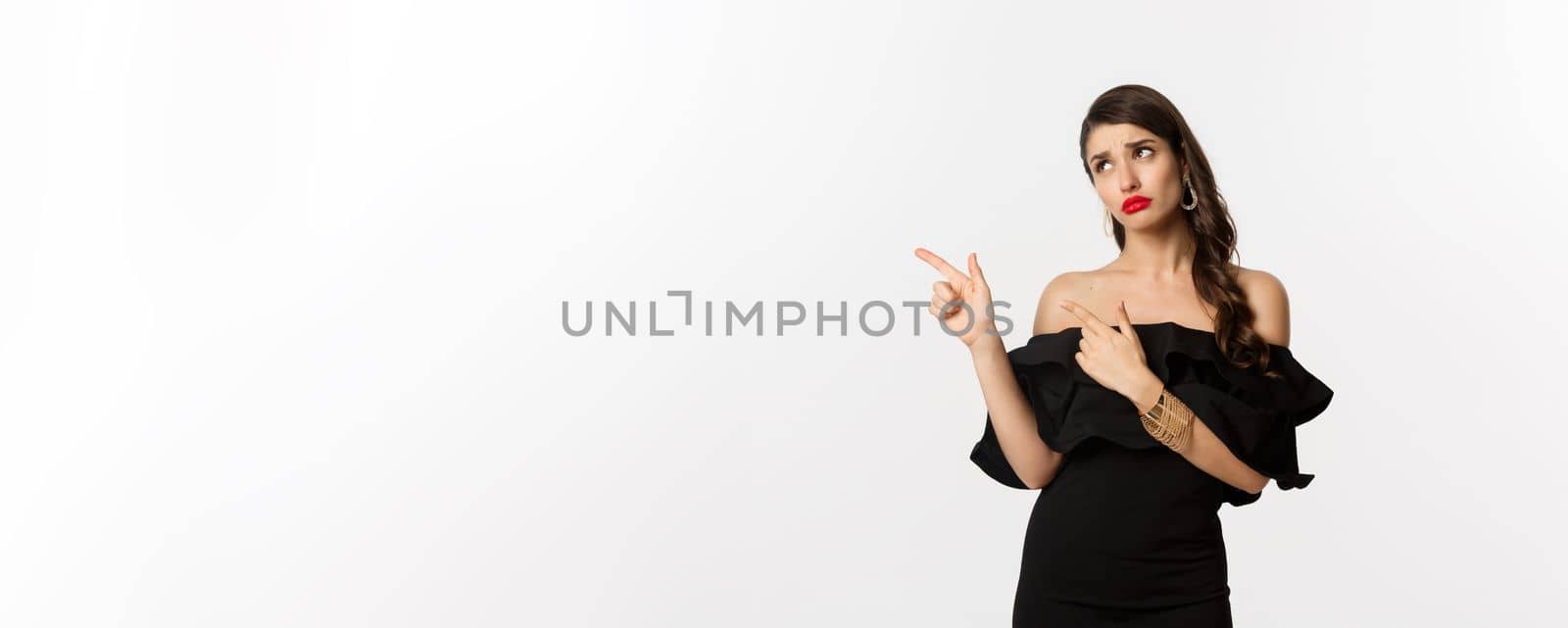 Fashion and beauty. Jealous glamour woman in black dress looking and pointing fingers left, sulking disappointed, white background.
