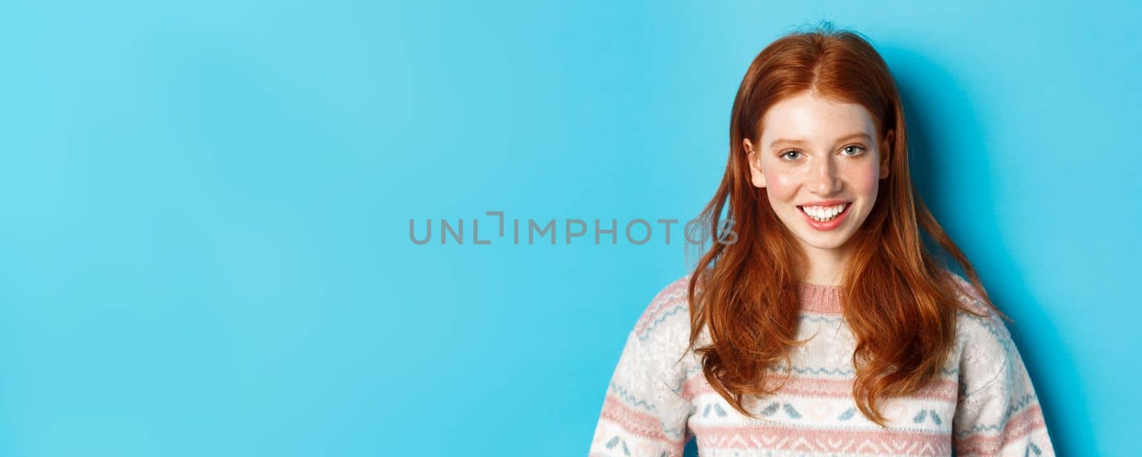 Close-up of cute redhead girl in sweater smiling happy at camera, standing against blue background by Benzoix