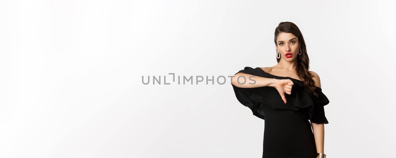 Fashion and beauty. Disappointed sassy woman in black dress, showing thumbs down, dislike something bad, judging over white background.