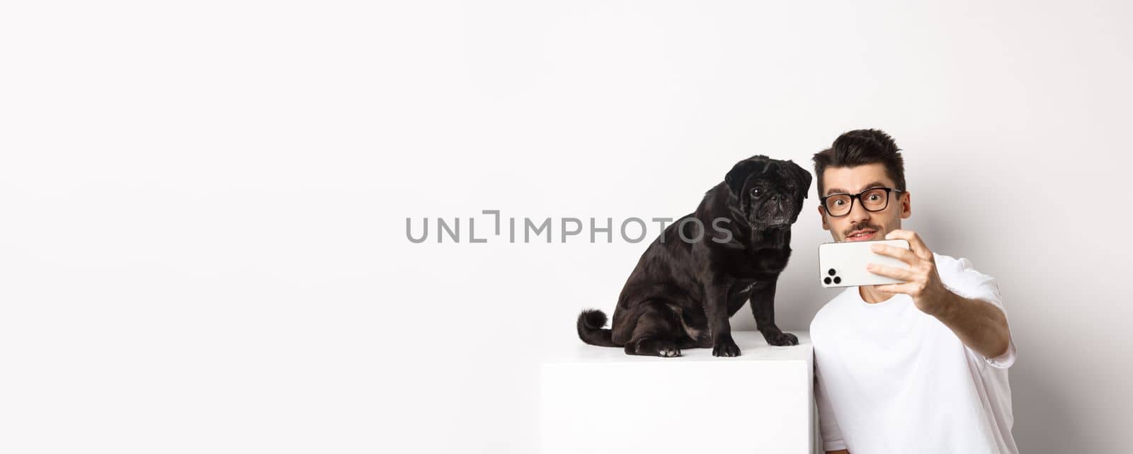 Image of handsome young man taking selfie with cute black dog on smartphone, posing with pug over white background.